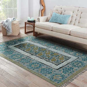 May include: A rectangular area rug with a floral design in shades of teal, light blue, and gold. The rug has a central floral pattern, a light blue border, and a fringe edge. The rug is placed on a hardwood floor.