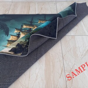 May include: A rolled-up rug featuring a detailed illustration of a tall ship sailing on the ocean. The design includes a palm tree, rocky cliffs, and a dark blue sky. The rug's underside is black with a white dotted pattern. The word "SAMPLE" is printed in red.