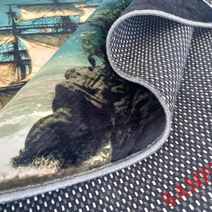 May include: A rolled-up rug featuring a detailed illustration of a tall ship sailing on the ocean. The rug's design includes a rocky coastline and waves. The underside of the rug is black with a white grid pattern and the word "SAMPLE" in red.