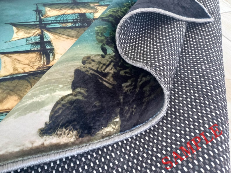 May include: A rolled-up rug featuring a detailed illustration of a tall ship sailing on the ocean. The rug's design includes a rocky coastline and waves. The underside of the rug is black with a white grid pattern and the word "SAMPLE" in red.