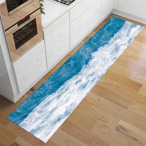 May include: A long, rectangular kitchen rug with a vibrant ocean wave design. The rug features a dynamic pattern of white foamy waves against a deep blue background. The rug is placed on a wooden floor, adding a touch of coastal decor.