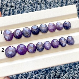 May include: Two rows of oval-cut gemstones in shades of purple and blue are displayed on a white surface. The gemstones have a faceted cut, reflecting light. The arrangement is numbered 1 and 2, with each row containing several stones.