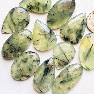 May include: A selection of polished prehnite gemstones in various shapes, including oval and teardrop. The translucent stones are pale green with black inclusions, and some have a yellowish hue. The gemstones are displayed on a light surface.