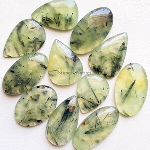 May include: A selection of polished prehnite gemstones in diverse shapes, such as teardrops and ovals. The translucent stones exhibit a pale green colour with black inclusions and a glossy finish. The gemstones are arranged on a white surface.