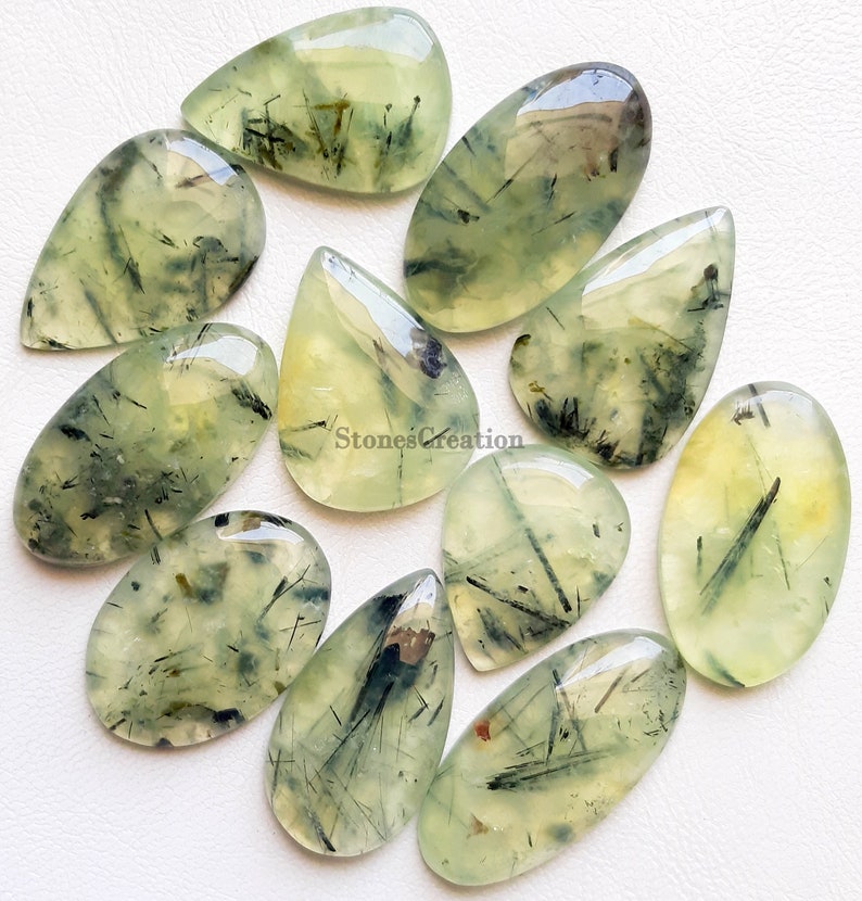 May include: A selection of polished prehnite gemstones in diverse shapes, such as teardrops and ovals. The translucent stones exhibit a pale green colour with black inclusions and a glossy finish. The gemstones are arranged on a white surface.