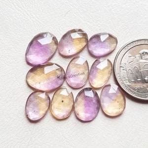 Pu&ograve; includere: Una collezione di dieci pietre preziose ovali sfaccettate, ametista e citrino. Le pietre sono un mix di viola chiaro e giallo, alcune con un gradiente di colore. Le pietre sono disposte in uno schema casuale su una superficie bianca.
