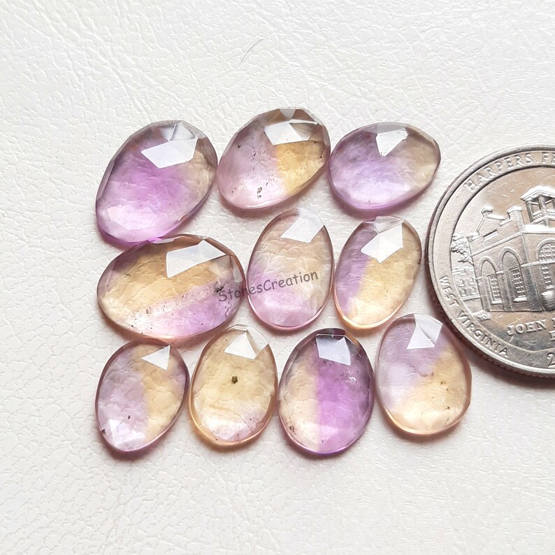 Pu&ograve; includere: Una collezione di dieci pietre preziose ovali sfaccettate, ametista e citrino. Le pietre sono un mix di viola chiaro e giallo, alcune con un gradiente di colore. Le pietre sono disposte in uno schema casuale su una superficie bianca.