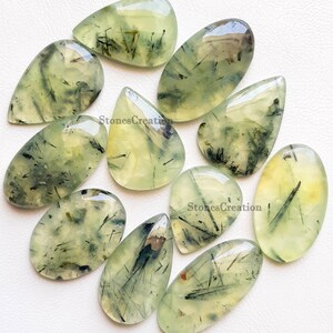 May include: A selection of polished prehnite gemstones. The stones are translucent with a pale green colour and feature dark, needle-like inclusions. The shapes vary, including teardrop and oval forms. The text "StonesCreation" is visible.