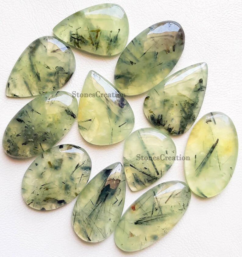 May include: A selection of polished prehnite gemstones. The stones are translucent with a pale green colour and feature dark, needle-like inclusions. The shapes vary, including teardrop and oval forms. The text "StonesCreation" is visible.
