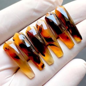 Op de afbeelding: Een set van zes lange, doodskistvormige acrylnagels. De nagels zijn een mix van amber, bruin en transparant, met een facetachtig, edelsteenachtig uiterlijk. De nagels zijn gerangschikt op een wit oppervlak.