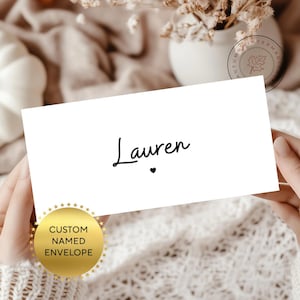 May include: A white envelope with the name "Lauren" in black script, with a small heart below the name. A gold sticker on the bottom left corner reads "CUSTOM NAMED ENVELOPE". The background is a soft, neutral color.