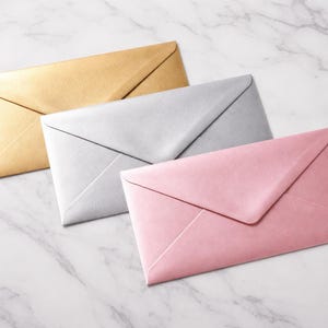 May include: Three colorful envelopes in gold, silver, and pink are arranged on a marble surface. The envelopes are closed and have a classic rectangular shape, suitable for invitations or letters. The image is well-lit, highlighting the colors.