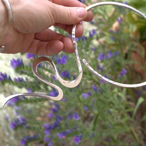 May include: A silver necklace with a distinctive, flowing design. The necklace has a hammered texture and an open, curved shape. The background features purple flowers and greenery, providing a natural contrast to the shiny metal.