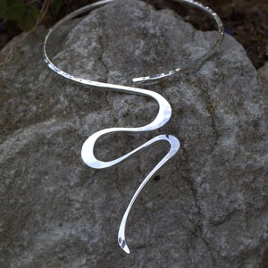 May include: A silver necklace with a hammered texture. The necklace features a curved, open design that encircles the neck and flows into a serpentine shape. The jewellery is displayed on a grey stone surface.