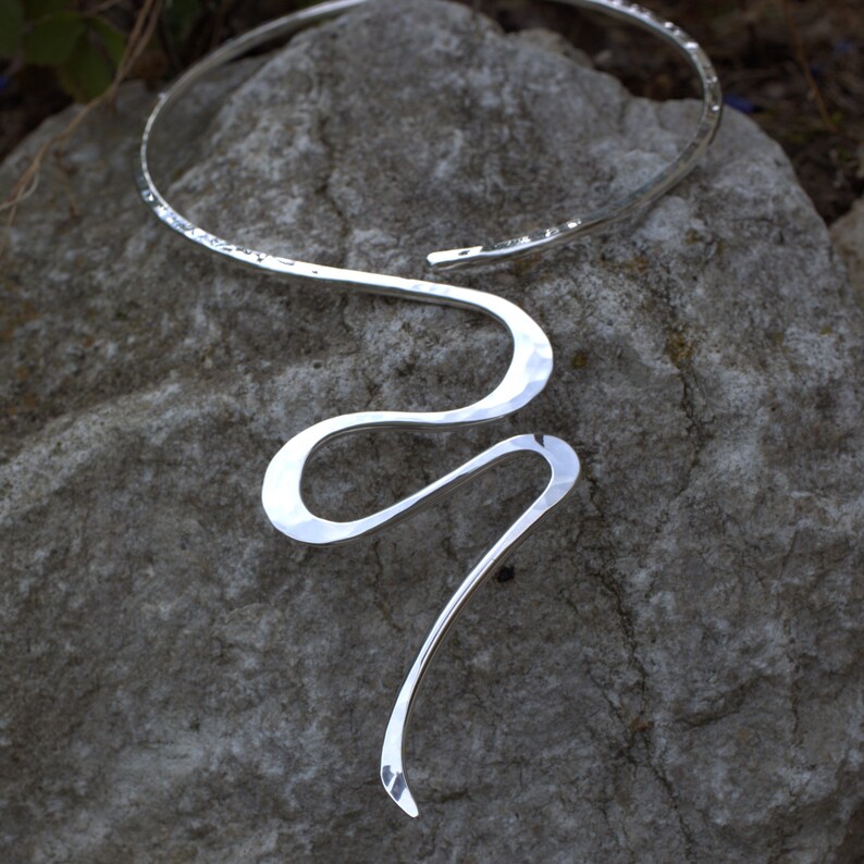 May include: A silver necklace with a hammered texture. The necklace features a curved, open design that encircles the neck and flows into a serpentine shape. The jewellery is displayed on a grey stone surface.