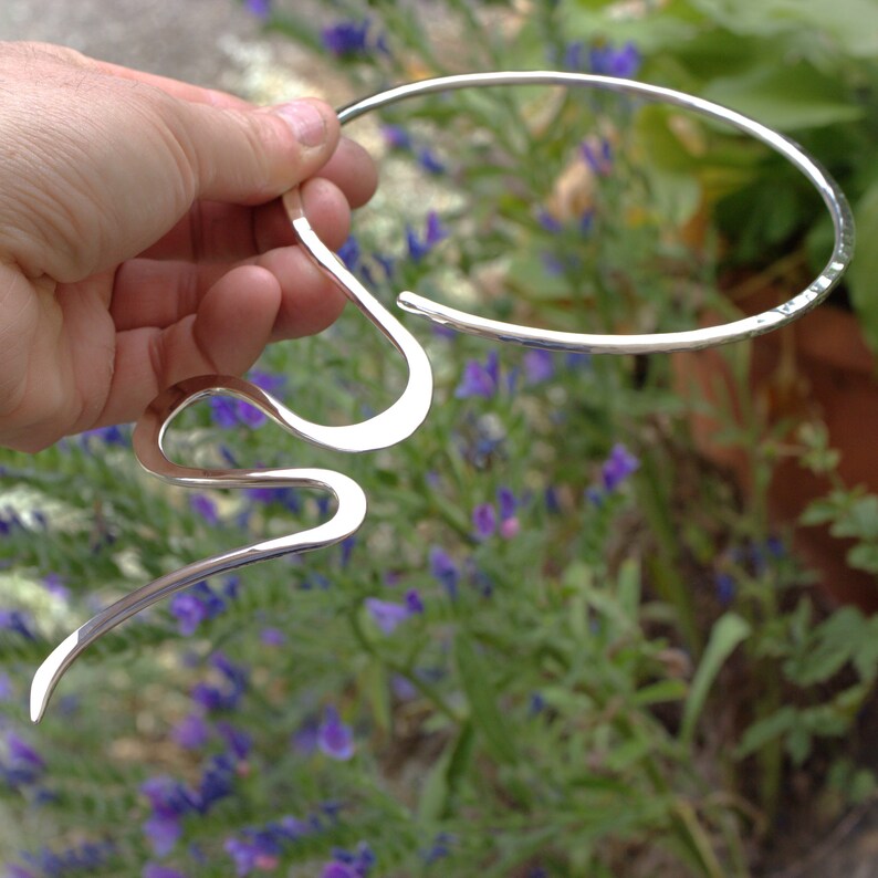 May include: A silver necklace with a distinctive, flowing design. The necklace features a curved, open-ended collar and a long, wavy pendant. The metal has a polished finish, reflecting light. The background shows green foliage and purple flowers.
