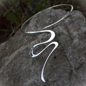 May include: A silver-coloured, open-ended necklace with a hammered texture on the upper section. The necklace has a flowing, abstract design with a curved, elongated shape. The jewellery is displayed on a grey stone surface.