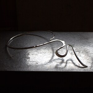 May include: A silver necklace with an abstract, flowing design. The necklace has a curved, open collar that flows into an artistic, asymmetrical shape. The metal has a hammered texture, adding visual interest to the jewellery. The necklace is made of silver.
