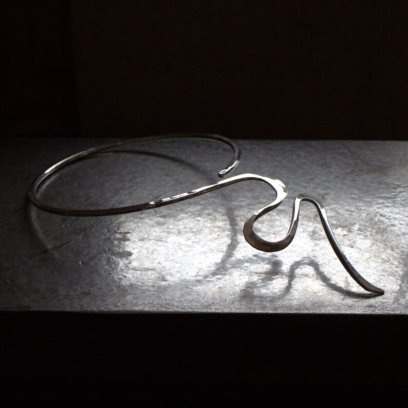 May include: A silver necklace with an abstract, flowing design. The necklace has a curved, open collar that flows into an artistic, asymmetrical shape. The metal has a hammered texture, adding visual interest to the jewellery. The necklace is made of silver.