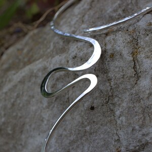 May include: A silver-coloured, hammered metal necklace with a flowing, open design. The necklace curves in a wave-like pattern, with a textured surface. The jewellery is displayed on a grey stone surface.