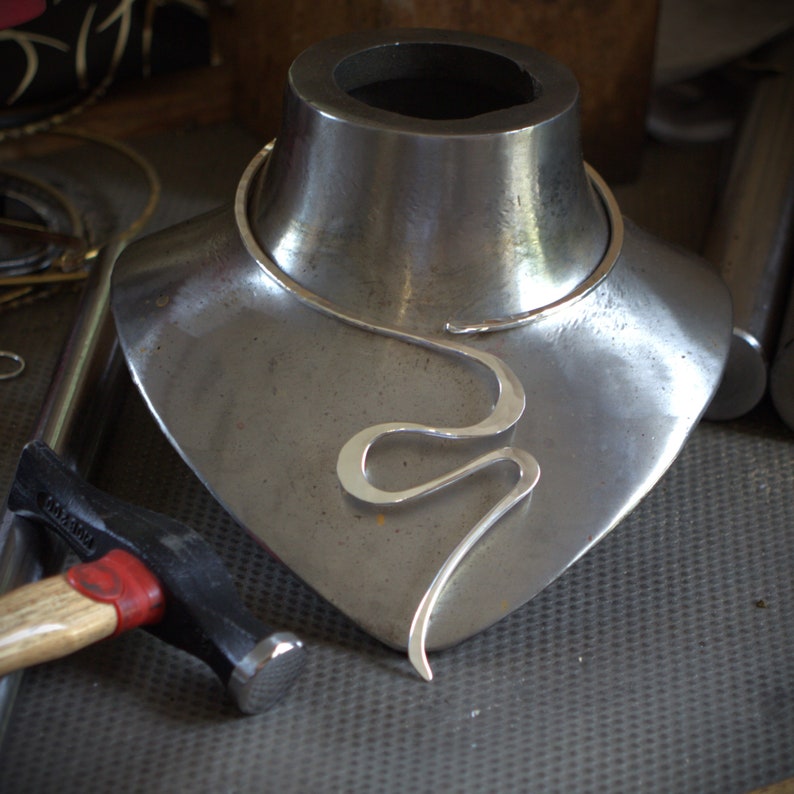 May include: A silver necklace with a flowing, abstract design is displayed on a metal bust. The necklace features a simple choker and a stylized, curved pendant. A hammer rests in the foreground, suggesting jewelry making.