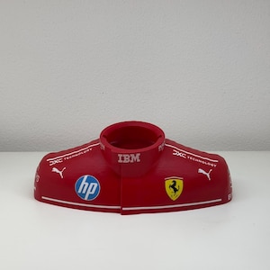 Könnte beinhalten: Rotes und weißes Modell eines Rennfahrerhelms mit Logos für IBM, HP, Puma und Ferrari.