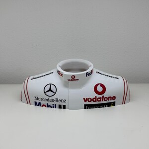 Può includere: Parte bianca di auto da corsa con dettagli rossi e neri. Presenta il logo Mercedes-Benz, il marchio Vodafone e Bridgestone. La parte ha un'apertura circolare nella parte superiore.