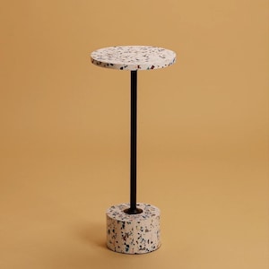 Könnte beinhalten: Ein kleiner, runder, weißer Beistelltisch mit schwarzen Beinen und einer Terrazzo-Oberfläche. Der Tisch steht auf einem hellbraunen Hintergrund.