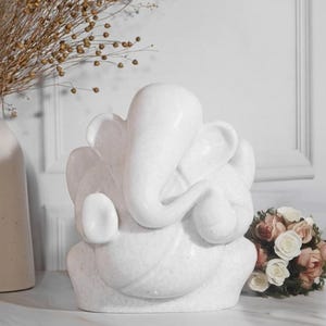 Puede incluir: Una escultura de mármol blanco del dios hindú Ganesha, representado con cabeza de elefante y múltiples brazos. La escultura tiene curvas suaves y está sobre una superficie de mármol. Un ramo de rosas y flores secas está en el fondo.
