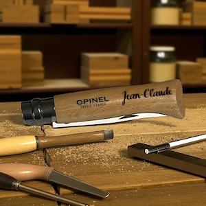 Peut inclure: Un couteau pliant Opinel à manche en bois avec le nom "Jean Claude" gravé sur le manche. Le couteau est ouvert, révélant la lame. D'autres outils de menuiserie sont visibles sur une surface en bois.