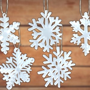 Puede incluir: Una cadena de luces de copo de nieve blanco con cinco copos de nieve colgando de un fondo de madera marrón.