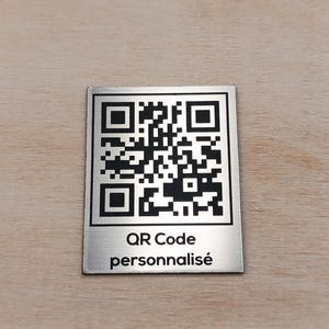 Puede incluir: Una placa rectangular de metal plateado con un código QR impreso en negro. Debajo del código QR, el texto dice "QR Code" en la primera línea y "personnalisé" en la segunda línea con una fuente negra sin remates.