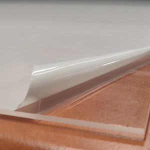 Könnte beinhalten: Eine transparente Acrylplatte mit einer Schutzfolie, die abgezogen wird. Die Platte liegt auf einer braunen Oberfläche.
