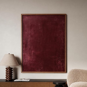 May include: A large, rectangular abstract painting with a deep burgundy color, framed in a dark wood. The artwork hangs on a neutral-toned wall, complemented by a wooden cabinet, a lamp with a beige shade, and a cozy armchair.