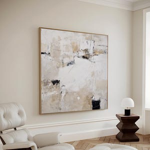 Puede incluir: Obra de arte abstracta sobre lienzo en tonos neutros de blanco, beige y negro, enmarcada en dorado. La obra cuelga sobre un conjunto de sillón y otomana de cuero blanco con base de madera. También hay una pequeña mesa auxiliar con una lámpara.