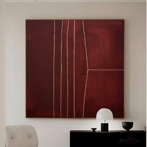 Puede incluir: Una gran pintura abstracta enmarcada en tonos burdeos con líneas doradas verticales y horizontales. La obra de arte cuelga sobre un mueble negro con una lámpara blanca y un cuenco negro. Una silla y un otomán blancos están en primer plano. El texto "CURIOSITY ES ART | HIGH QUALITY CANVAS" está en la parte inferior.