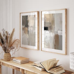 Puede incluir: Dos impresiones artísticas abstractas enmarcadas en tonos neutros cuelgan sobre una consola de madera. La obra presenta pinceladas beige, grises y negras. Un jarrón con flores secas, libros y objetos decorativos completan la escena. El texto "CURIOSITY ES ART | PRINTABLE WALL ART" es visible.