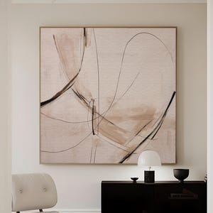 Puede incluir: Lienzo de arte abstracto con pinceladas beige y marrón y líneas negras. La obra de arte está enmarcada en un tono dorado y cuelga sobre una moderna silla y otomana. El texto "CURIOSITY ES ART | HIGH QUALITY CANVAS" está en la parte inferior.