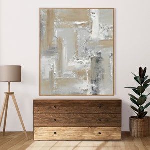 Puede incluir: Una pintura abstracta enmarcada con un esquema de color beige, gris y blanco. La pintura está colgada en una pared blanca sobre una cómoda de madera con tres cajones.