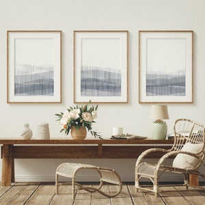 Puede incluir: Tres impresiones de arte abstracto enmarcadas con un esquema de color azul y blanco cuelgan de una pared blanca sobre una mesa de madera con una silla de mimbre y un taburete.
