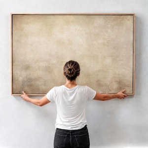 Puede incluir: Un gran lienzo rectangular enmarcado con un diseño abstracto texturizado neutro en tonos beige y marrón. La obra de arte está sostenida por una persona que lleva una camiseta blanca y vaqueros negros.