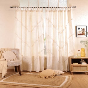Pode incluir: Cortinas brancas de algodão com um padrão de diamante e acabamento em franjas. As cortinas estão penduradas em uma haste preta e estão parcialmente abertas, revelando uma janela com vista para uma parede branca.