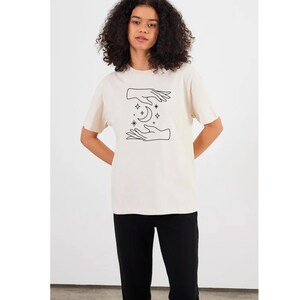 May include: A cream colored t-shirt with a black line drawing of two hands cupping a crescent moon with stars.