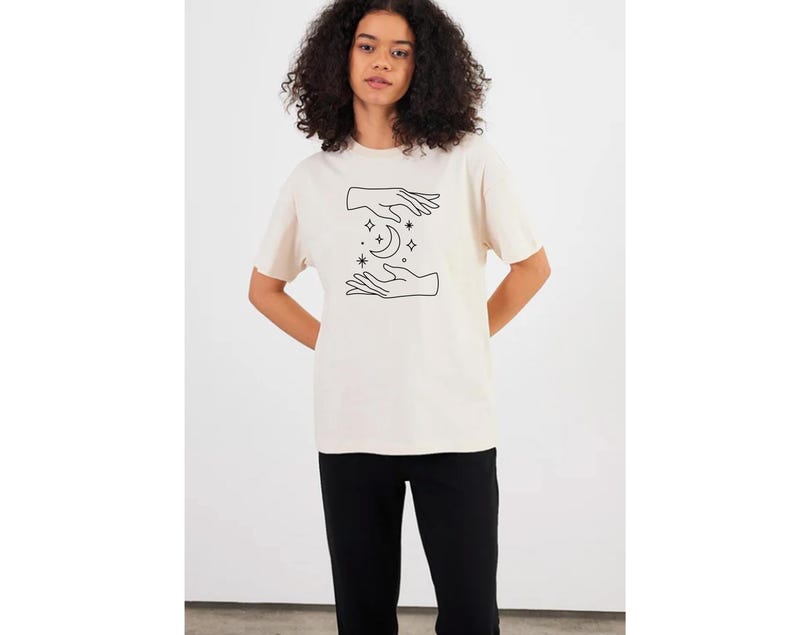 May include: A cream colored t-shirt with a black line drawing of two hands cupping a crescent moon with stars.