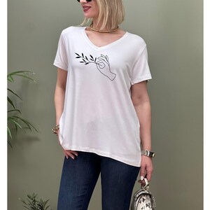 May include: White oversized v-neck t-shirt with a black line drawing of a hand holding a branch with leaves.