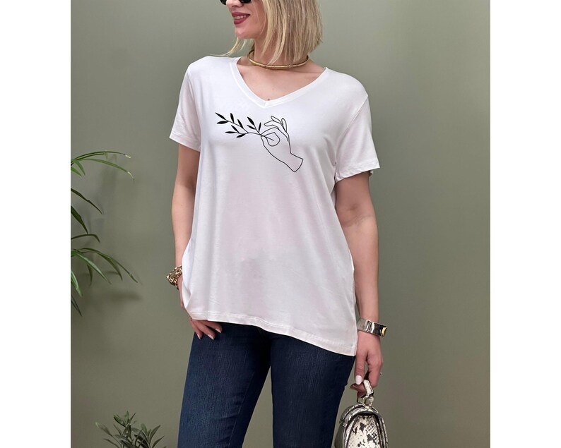 May include: White oversized v-neck t-shirt with a black line drawing of a hand holding a branch with leaves.