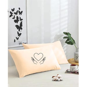 May include: Two peach-colored pillowcases with a black line drawing of hands holding a heart on one pillowcase.