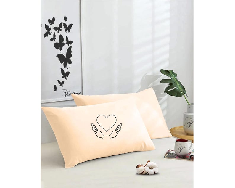 May include: Two peach-colored pillowcases with a black line drawing of hands holding a heart on one pillowcase.