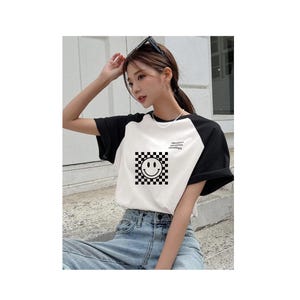 Może przedstawiać: Biało-czarna koszulka typu raglan z nadrukiem uśmiechniętej buźki w czarno-białą kratkę. Na koszulce widnieje napis "Vacation Mode: Activated".