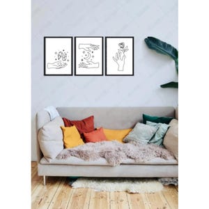May include: Three black framed prints with line drawings of hands holding a crescent moon, a flower, and a star. The prints are hanging on a white wall above a light gray couch with colorful throw pillows.